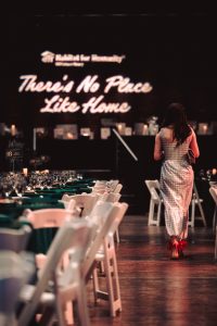 Woman in a gingham dress walking towards "there's no place like home" projected on the wall.